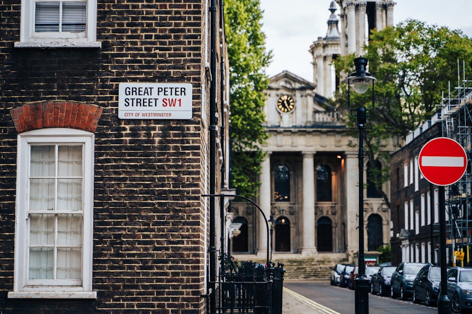 A street corner featuring a brick building with a white-framed window and an street sign indicating Great Peter Street SW1 in the City of Westminster. In the background, there is an ornate classical-s